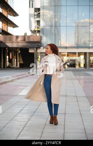 Una donna latina con capelli ricci e caratteristiche miste cammina con sicurezza attraverso un paesaggio urbano moderno indossa un cappotto elegante, portando con sé un caffè e una linguetta Foto Stock