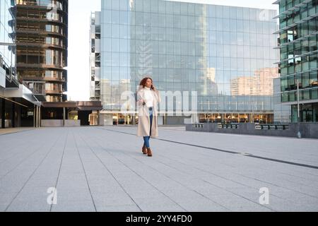 Una donna latina sicura di sé con i capelli ricci cammina attraverso un paesaggio urbano moderno, tenendo in mano un computer portatile gli edifici di vetro e l'ambiente urbano riflettono un professionista Foto Stock