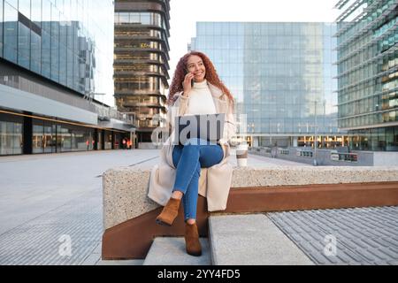 Una donna latina con i capelli ricci siede su una panchina della città, sorridendo mentre parla al telefono e lavora su un portatile i moderni edifici in vetro creano un ambiente contemporaneo Foto Stock