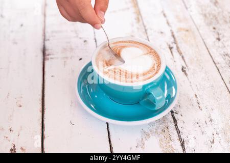 Primo piano di una mano che mescola un cappuccino con delicata latte art a forma di cuore in una tazza blu. L'ambiente e' un rustico tavolo di legno, che mette in risalto un co Foto Stock