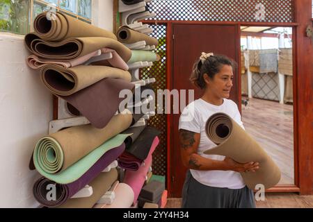 Una donna in uno studio di yoga tiene un tappetino arrotolato, organizzando diversi tappetini colorati sugli scaffali a parete. Lo spazio presenta pavimenti in legno e pareti a motivi geometrici. Foto Stock