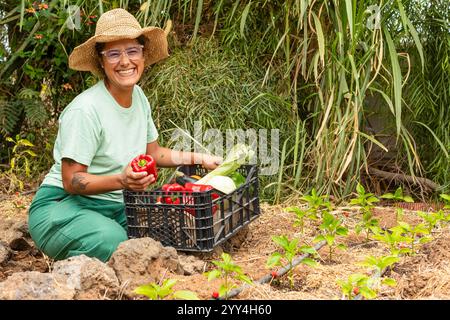 Una donna che indossa un cappello sorride mentre raccoglie verdure fresche dal suo giardino le piante giovani crescono con l'aiuto di un sistema di irrigazione a goccia visibile Foto Stock