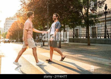 Una felice coppia gay che si tiene per mano mentre attraversano una strada urbana illuminata dal sole, sorridendo e godendo una giornata calda Foto Stock