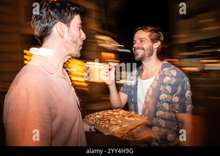 Una felice coppia gay condivide una fetta di pizza mentre cammina e ridi insieme in un vivace ambiente cittadino di notte, catturando una roma spontanea Foto Stock