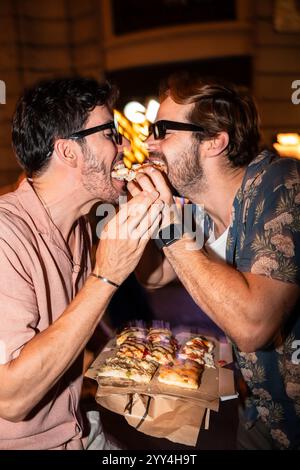 Un'affascinante scena di una coppia gay che condivide una fetta di pizza, nutrendosi amorevolmente in un vivace ambiente all'aperto di notte Foto Stock