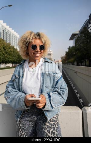 Una giovane donna domenicana afro dai capelli biondi ricci, con occhiali da sole e giacca in denim, sorride brillantemente tenendo in mano uno smartphone in un moderno urb Foto Stock