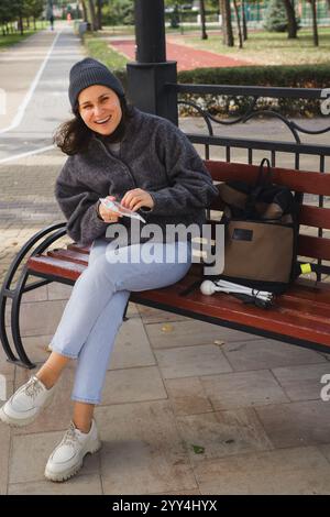 Una donna allegra con problemi visivi siede su una panchina del parco, sorridendo tiene uno spuntino e la sua borsa con un bastone bianco riposa accanto a lei, a simboleggiare Foto Stock