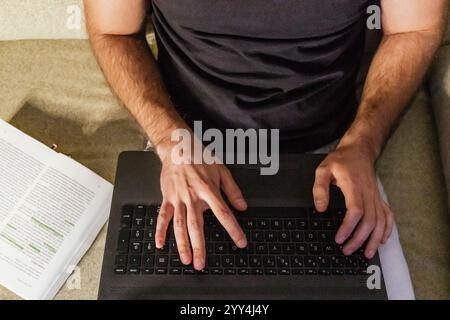 Vista dall'alto di una persona che digita su un notebook, lavora su attività di social media da casa Un documento stampato si trova nelle vicinanze, suggerendo ricerche e pianificazione Foto Stock