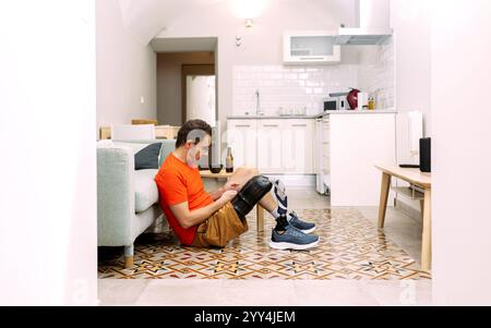 Un uomo di mezza età con una gamba protesica siede su un pavimento piastrellato in una cucina luminosa e moderna indossa abiti casual e sembra rilassato, ritraendo sempre Foto Stock