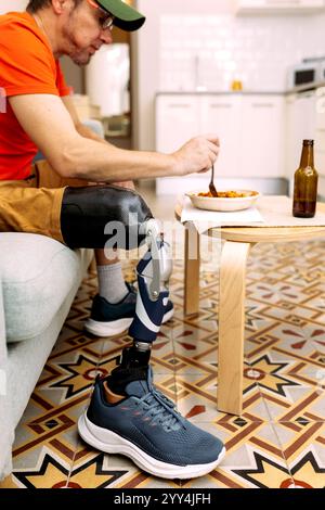 Un uomo che indossa una camicia arancione e un berretto è seduto su un divano con la gamba protesica visibile, sta mangiando da una ciotola su un tavolo di legno in un kitc moderno Foto Stock