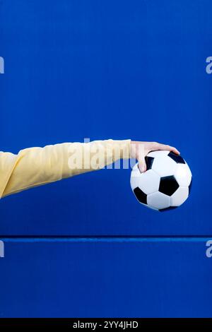 Un'immagine minimalista che mostra una mano in una manica gialla che regge un classico pallone da calcio bianco e nero su uno sfondo blu vivace, sottolineando la S. Foto Stock