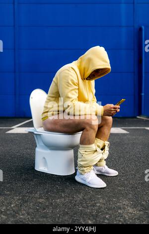 Un uomo con cappuccio giallo siede in un bagno all'aperto, concentrandosi sul suo smartphone. Il netto contrasto contro il muro blu crea un umorismo e una strikin Foto Stock