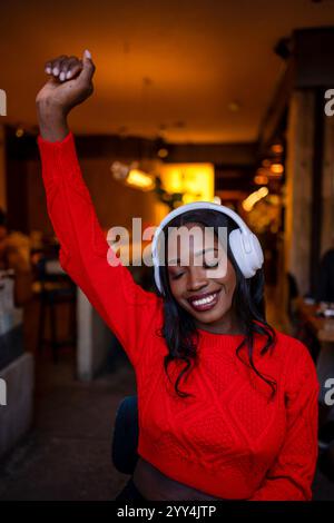 Un cliente soddisfatto che indossa un maglione rosso ascolta la musica con le cuffie e solleva il braccio in un bar Foto Stock