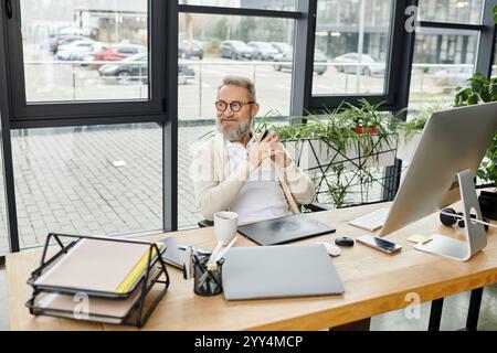 Un uomo bello e maturo siede comodamente ad una scrivania, concentrandosi sul lavoro, circondato dal verde. Foto Stock