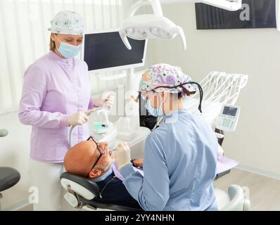 Dentista che indossa lenti d'ingrandimento che esegue l'esame dentistico su un paziente di sesso maschile anziano mentre l'infermiera la assiste in una moderna clinica odontoiatrica Foto Stock