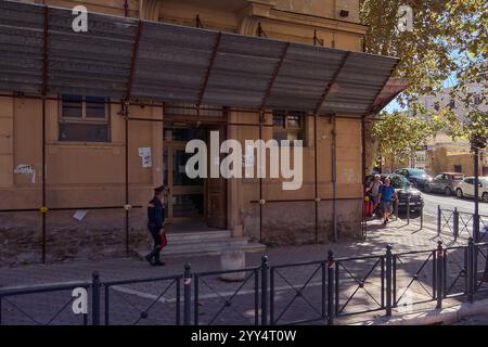 Civitavecchia, Italia - 19 dicembre 2024: Vecchio edificio con facciata intemprata e tenda metallica, con persona in uniforme che cammina e auto parcheggiate lungo il Foto Stock