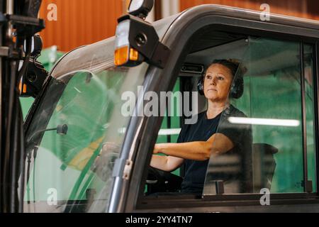 Lavoratrice matura che indossa le cuffie wireless mentre è seduta sul carrello elevatore in fabbrica Foto Stock