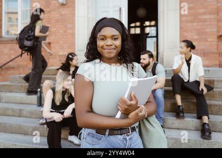 Ritratto di una giovane donna sorridente che tiene un libro mentre si trova al campus universitario Foto Stock