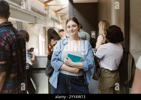 Ritratto di una giovane donna sorridente che tiene un libro mentre si trova vicino agli amici nel corridoio del college Foto Stock