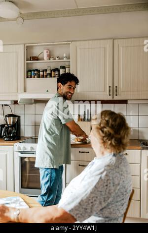 Vista laterale dell'infermiera sorridente che prepara la colazione mentre parla con una donna anziana in cucina a casa Foto Stock