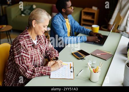 Vista laterale di una dipendente matura esperta che pianifica attività mensili scrivere su un tavolo del calendario mentre si siede alla scrivania in ufficio, copiare spazio Foto Stock