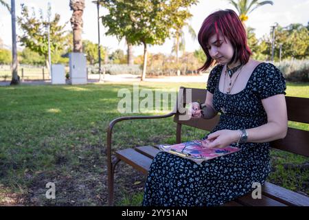 La giovane donna seduta su una panchina del parco sta infilando perline in cotone per rendere disponibile un bracciale Foto Stock