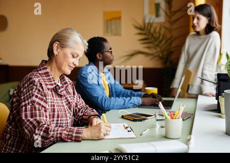 Vista laterale di una dipendente matura che scrive nella tabella del calendario pianificando le attività mensili mentre si è seduti alla scrivania in ufficio, spazio per le copie Foto Stock