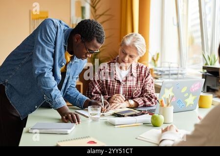 Giovane uomo di colore con occhiali che lascia una nota su carta per un collega anziano su come utilizzare correttamente il notebook assistendo la donna nello sviluppo professionale, mentre lavora alla scrivania in ufficio Foto Stock