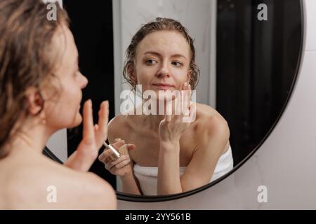 donna che applica cosmetici di fronte allo specchio in bagno, cura della pelle del viso, Foto Stock