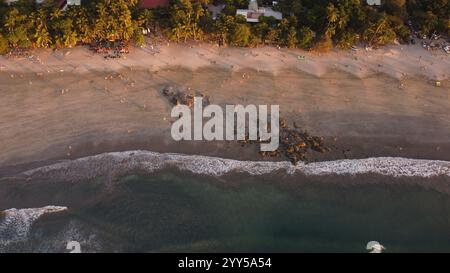 Vista aerea di una spiaggia tropicale al tramonto con palme, costa rocciosa e visitatori sparsi che si godono il tranquillo paesaggio costiero. Foto Stock