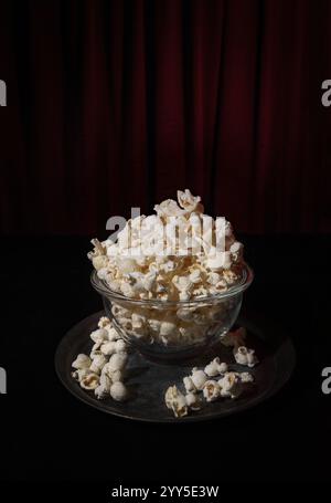 Un'unica ciotola di popcorn appena sfornati collocati su uno sfondo scuro con sfumature rosse che ricordano una tenda teatrale, evocando un atmos cinematografico Foto Stock