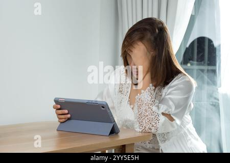 Una donna asiatica in un abito da notte in raso bianco prova dolore agli occhi e fatica mentre legge il suo programma di lavoro su un tablet, facendola stare al passo con lei Foto Stock