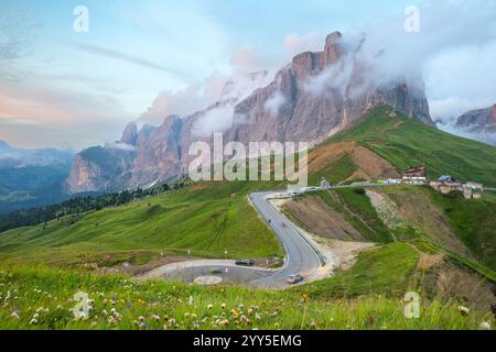 La montagna rocciosa circondata da nuvole con una strada curva e la collina scivolosa in primo piano al tramonto, passo Sella, Dolomiti, Italia Foto Stock