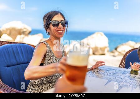 Donna che brinda con un bicchiere di birra in un ristorante sul mare a pirano, in slovenia, godendosi una giornata di sole piena di risate e relax Foto Stock