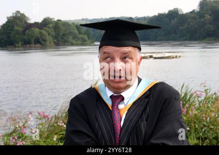 Uomo anziano con un'espressione buffa che indossa un cappello e un abito da graduazione in piedi sullo sfondo di un fiume panoramico. Foto Stock