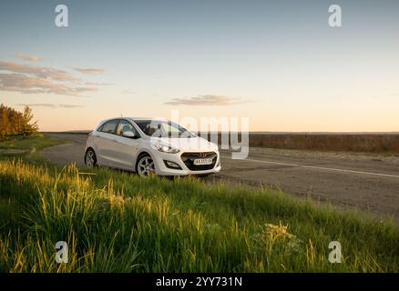 Hyundai i30 bianca parcheggiata lungo la strada, erba verde in primo piano. Berlina compatta coreana in zona rurale al tramonto. Foto Stock