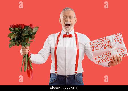 Uomo maturo scioccato con confezione regalo e bouquet di rose su sfondo rosso. Festa di San Valentino Foto Stock