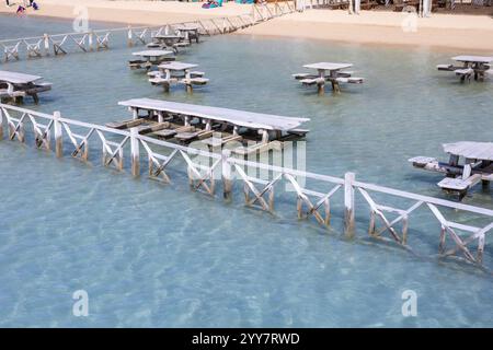 Una vista panoramica dei tavoli da picnic in legno in piedi in acque cristalline e poco profonde vicino a una spiaggia di sabbia Foto Stock