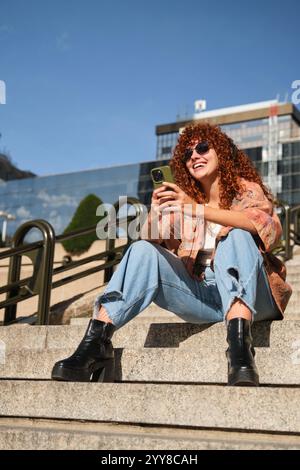 Donna rossa che usa il telefono e ascolta musica seduta sulle scale Foto Stock