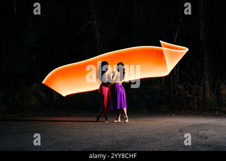 Due donne in piedi sotto una pista luminosa arancione in una foresta buia Foto Stock