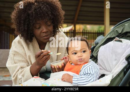 Adorabile madre che dà da mangiare al suo adorabile bambino nel passeggino del parco giochi Foto Stock
