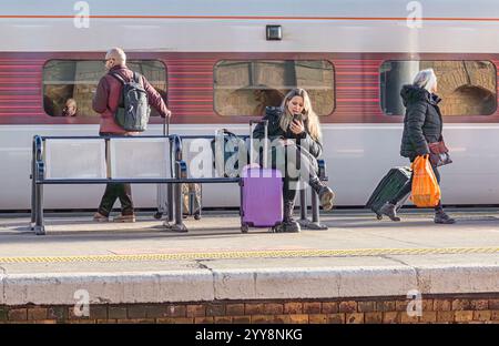 Tre passeggeri sono su una piattaforma della stazione ferroviaria. Uno è seduto su una panchina, uno sta in piedi e un altro cammina. Una carrozza ferroviaria è sullo sfondo. Foto Stock
