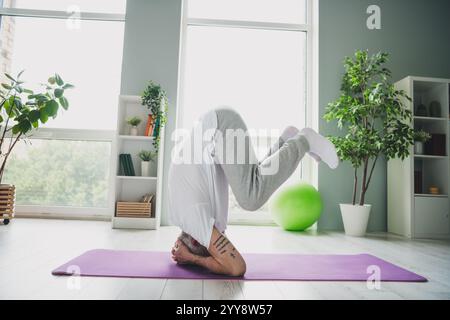 Uomo anziano che pratica il poggiatesta yoga su un tappetino viola in interni durante il giorno Foto Stock