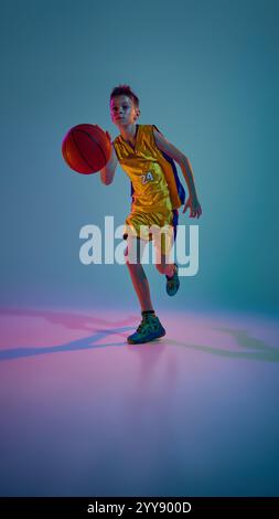 Incentrato sul gioco, atleta di basket junior, maglia gialla vestita, corsa in movimento con luce al neon sullo sfondo sfumato dello studio. Foto Stock