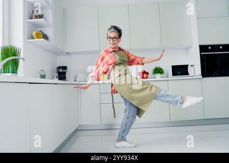 Gioiosa donna anziana che balla in una cucina moderna, si diverte a cucinare in casa, indossa abiti casual e grembiule Foto Stock