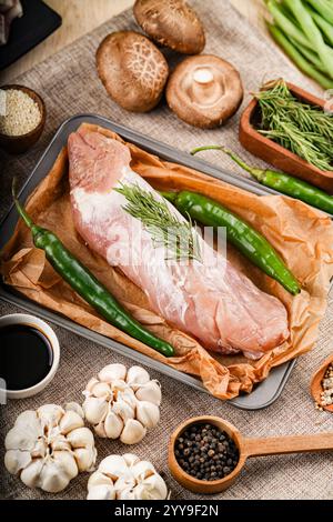 Spalla di maiale tagliata di fresco disposta su una tavola rustica di legno, circondata da verdure e spezie vivaci, pronta per un sostanzioso pasto casalingo. Foto Stock