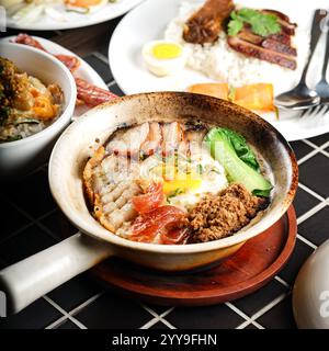 Tradizionale riso in pentola di argilla servito con una varietà di condimenti salati, tra cui pancetta di maiale croccante, salsiccia cinese, maiale tritato, uovo al sole e foglie Foto Stock
