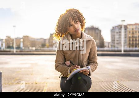 Pensiva bellezza donna latina che scrive appunti seduti sulla città con paesaggio urbano sullo sfondo in una giornata fredda Foto Stock