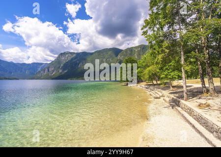 Le acque cristalline turchesi si infrangono dolcemente contro la spiaggia di ciottoli, incorniciate da alberi lussureggianti e dalle maestose alpi giulie sotto un vibrante cielo estivo Foto Stock