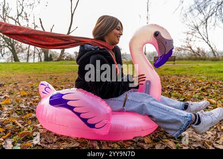 giovane bella donna che si diverte a cavalcare un fenicottero rosa gonfiabile in un parco pubblico in inverno Foto Stock
